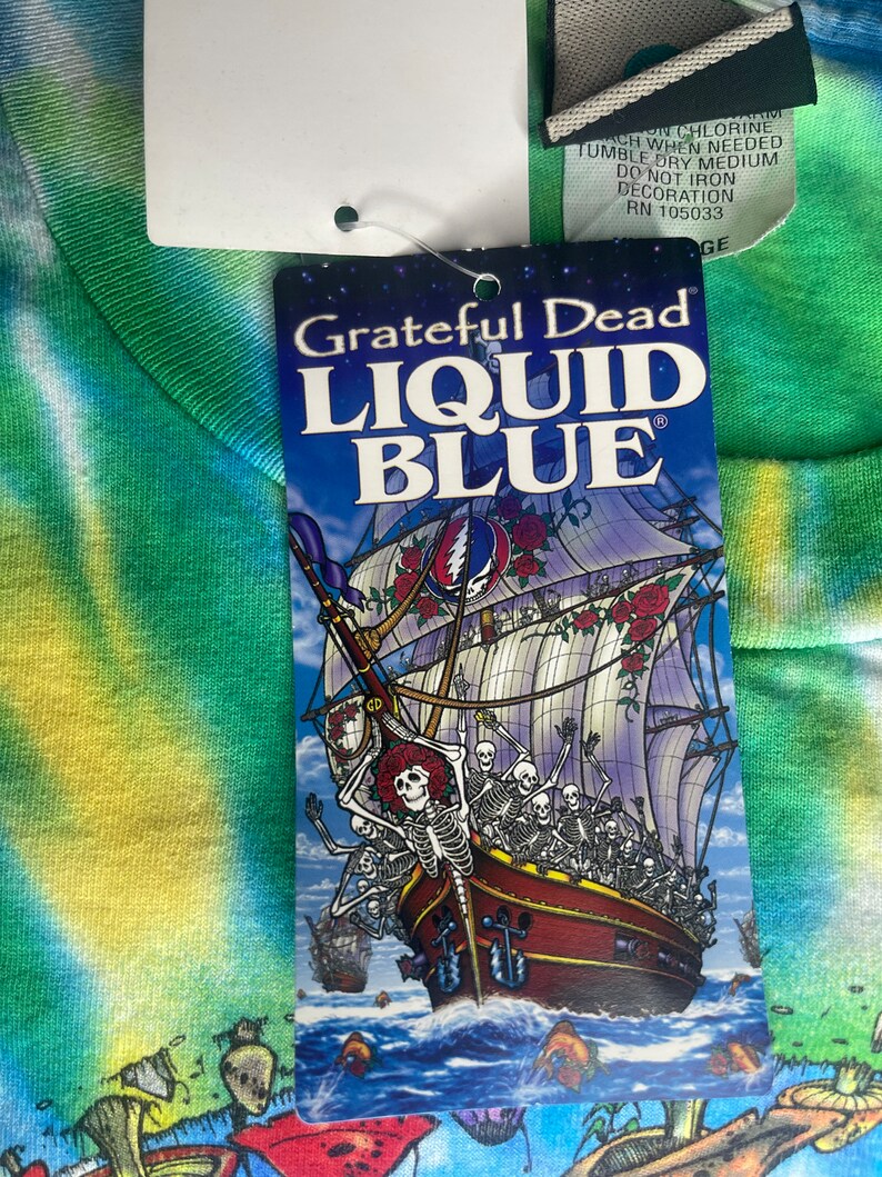 以下が含まれることがあります： スケルトンとバラの船が描かれたタグが付いたタイダイTシャツ。テキストは「Grateful Dead LIQUID BLUE」です。シャツは緑、青、黄色の模様です。