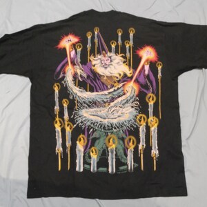 Op de afbeelding: Zwart T-shirt met een grafische print van een tovenaar met een witte baard en een paarse robe, omringd door kaarsen. De tovenaar houdt een staf vast en werpt een spreuk.