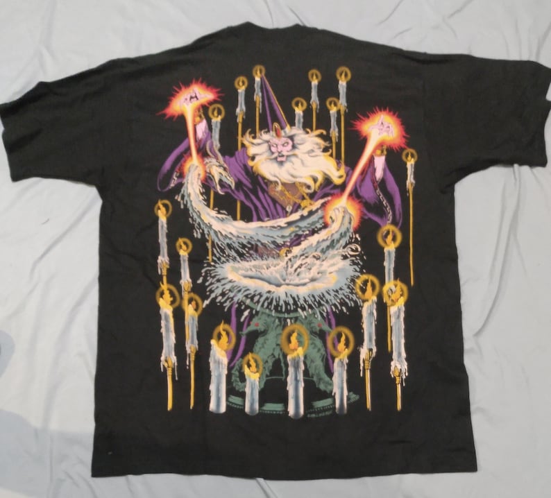 Op de afbeelding: Zwart T-shirt met een grafische print van een tovenaar met een witte baard en een paarse robe, omringd door kaarsen. De tovenaar houdt een staf vast en werpt een spreuk.