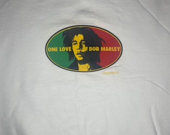 Puede incluir: Camiseta blanca con un gráfico colorido de Bob Marley. El gráfico presenta un fondo rojo, amarillo y verde con el texto "One Love" y "Bob Marley".