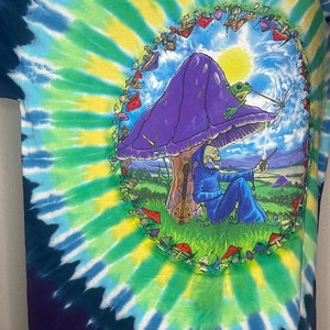 以下が含まれることがあります： 青、緑、黄色の鮮やかなスパイラル模様のタイダイTシャツ。紫色のキノコ、カエル、青いローブを着た骸骨の姿、ギターが描かれています。デザインはキノコの輪で囲まれています。