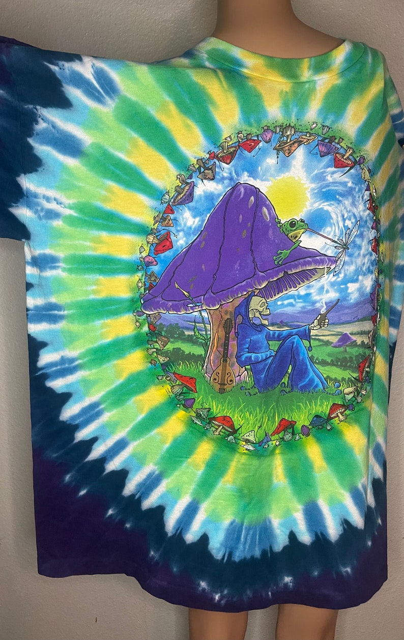 以下が含まれることがあります： 青、緑、黄色の鮮やかなスパイラル模様のタイダイTシャツ。紫色のキノコ、カエル、青いローブを着た骸骨の姿、ギターが描かれています。デザインはキノコの輪で囲まれています。
