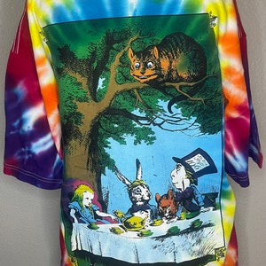 Op de afbeelding: Een tie-dye T-shirt met een kleurrijk ontwerp. De achterkant van het shirt heeft een afbeelding van een sc&egrave;ne uit Alice in Wonderland, met Alice, de Hoedenmaker, de Maartse Haas en de Cheshire Cat.