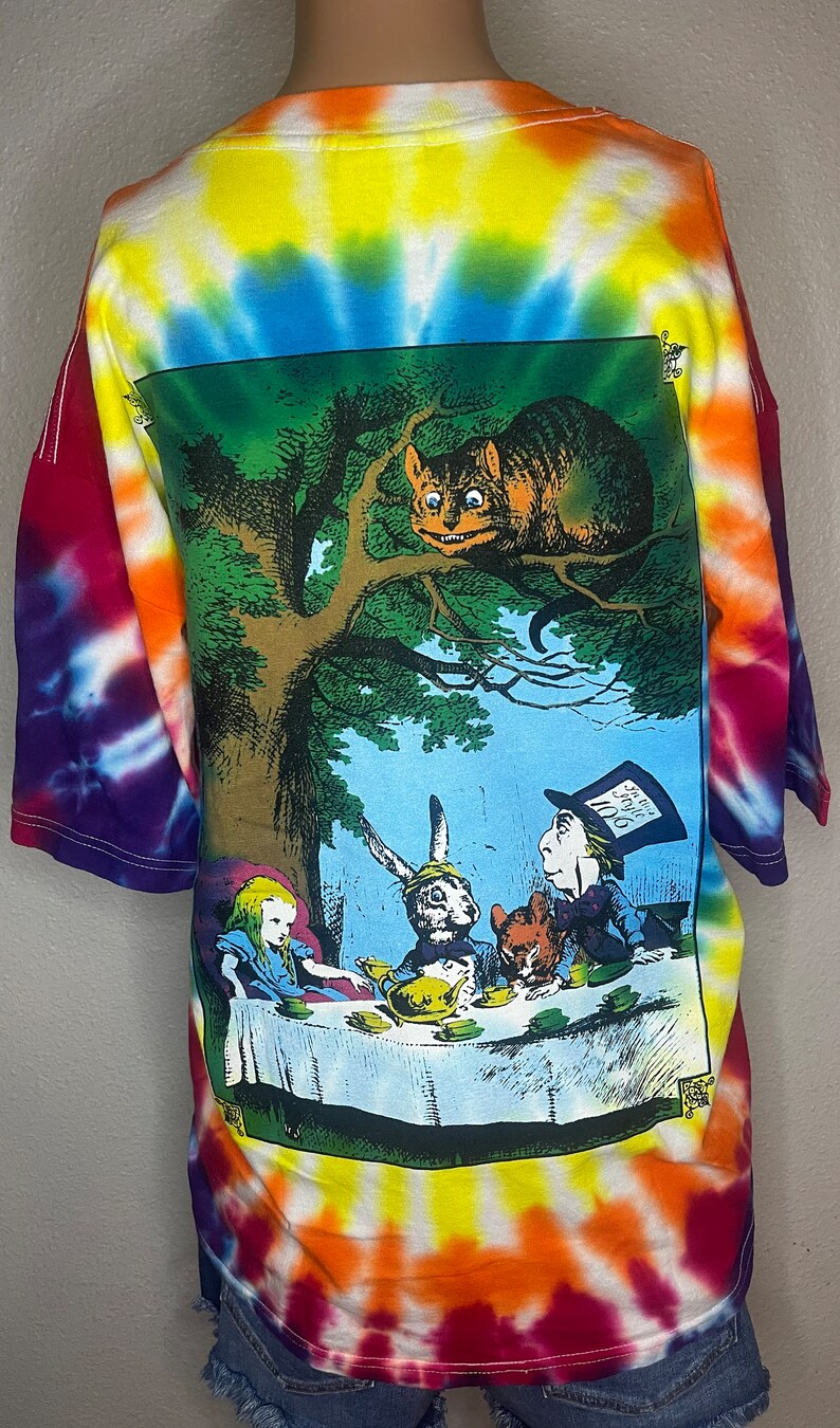 Op de afbeelding: Een tie-dye T-shirt met een kleurrijk ontwerp. De achterkant van het shirt heeft een afbeelding van een sc&egrave;ne uit Alice in Wonderland, met Alice, de Hoedenmaker, de Maartse Haas en de Cheshire Cat.