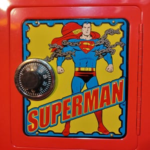 May include: Red metal safe with a Superman graphic on the front. The graphic features Superman in his classic costume, chained, with the word "SUPERMAN" in bold letters. A combination dial is on the left, and the text "Saving money is Powerful!" is below.