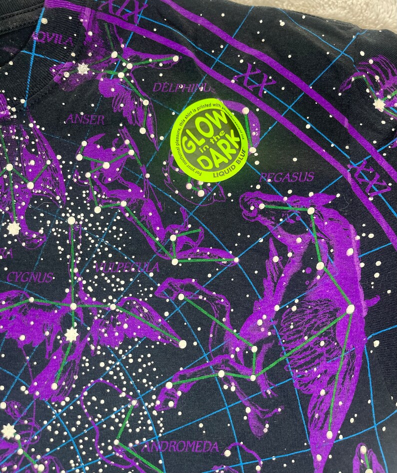 May include: Black t-shirt with a constellation map printed on it. The constellations are outlined in purple and the stars are white. The shirt has a glow-in-the-dark label that says "Glow in the Dark" with "Liquid Blue" printed below it.