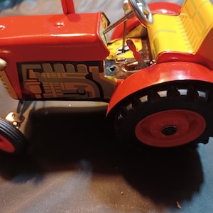 Pode incluir: Um trator de brinquedo vermelho e amarelo vintage com pneus pretos e um volante prateado. O trator apresenta um design detalhado do motor na lateral e um assento amarelo. O brinquedo é feito de metal e tem um apelo clássico e nostálgico.