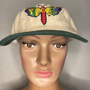 Könnte beinhalten: Eine beige Baseballkappe mit grünem Schirm und einem bunten Schmetterling, der auf der Vorderseite gestickt ist. Der Schmetterling hat das Wort "VOTE" auf seinen Flügeln.