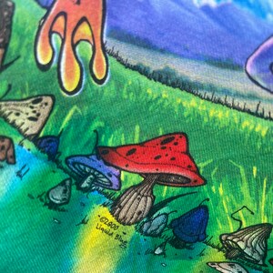 以下が含まれることがあります： カラフルなグラフィックTシャツのクローズアップ。キノコ、蝶、その他の気まぐれな要素の鮮やかなデザインが特徴です。デザインには、白い水玉模様の赤いキノコ、青いキノコ、そして「&copy;2000 Liquid Blue」の文字が含まれています。