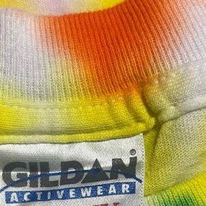 Op de afbeelding: Een wit label met zwarte tekst dat "Gildan Activewear Heavy Weight Cotton" en "100% Katoen Voorgekrompen Geassembleerd in Nicaragua van Canadese componenten" zegt. Het label is bevestigd aan een tie-dye shirt met oranje, gele, groene en blauwe kleuren.