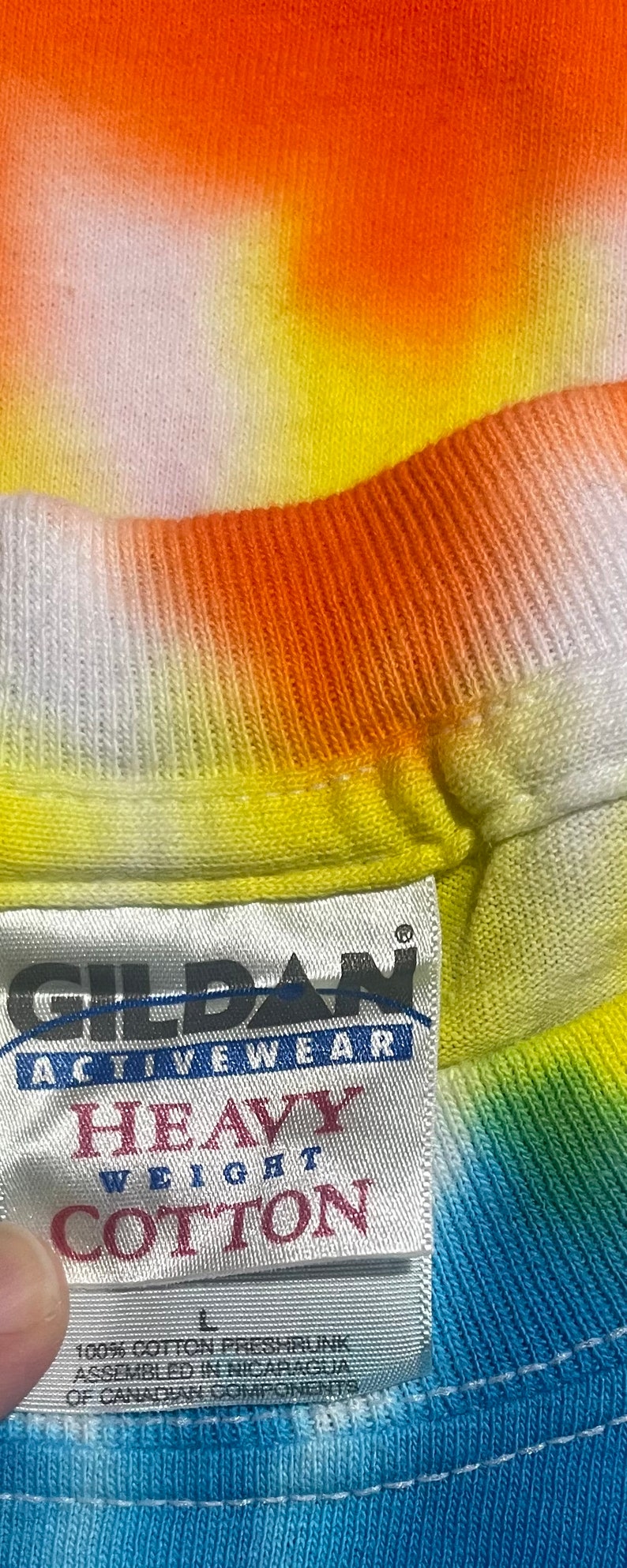 Op de afbeelding: Een wit label met zwarte tekst dat "Gildan Activewear Heavy Weight Cotton" en "100% Katoen Voorgekrompen Geassembleerd in Nicaragua van Canadese componenten" zegt. Het label is bevestigd aan een tie-dye shirt met oranje, gele, groene en blauwe kleuren.