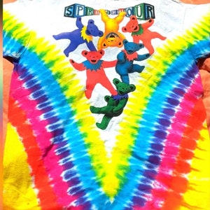 Puede incluir: Una camiseta blanca de tie-dye con un patr&oacute;n de arco&iacute;ris y un grupo de osos de dibujos animados coloridos colgando de la palabra "SPRINGTOUR" en letras de bloque.