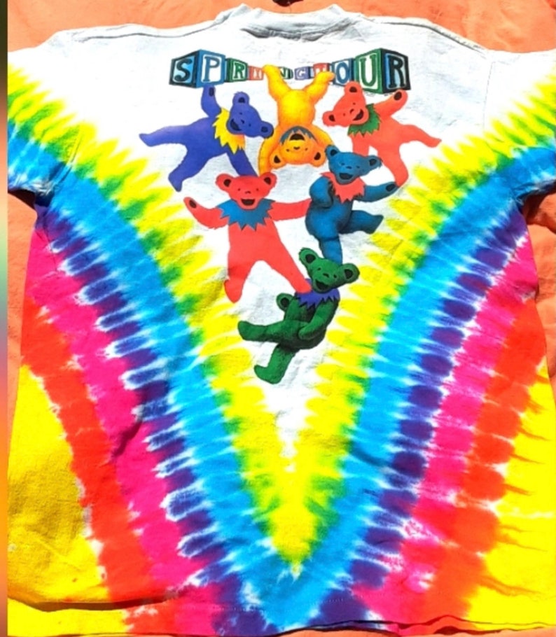 Puede incluir: Una camiseta blanca de tie-dye con un patr&oacute;n de arco&iacute;ris y un grupo de osos de dibujos animados coloridos colgando de la palabra "SPRINGTOUR" en letras de bloque.