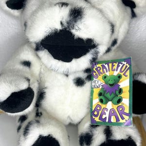May include: A white and black spotted plush bear with a black nose and mouth. The bear is wearing a purple and green bandana. The bear has a tag that says "Grateful Bear" with a green bear on it.