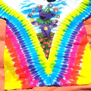 Puede incluir: Una camiseta tie-dye con un oso de dibujos animados morado que lleva un sombrero verde y hace malabares con bloques de colores. La camiseta tiene un patr&oacute;n tie-dye de arco&iacute;ris con amarillo, azul, rosa y rojo.