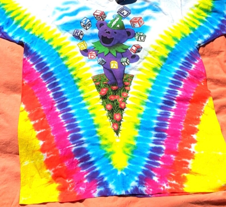 Puede incluir: Una camiseta tie-dye con un oso de dibujos animados morado que lleva un sombrero verde y hace malabares con bloques de colores. La camiseta tiene un patr&oacute;n tie-dye de arco&iacute;ris con amarillo, azul, rosa y rojo.