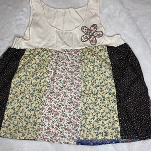 May include: A white sleeveless top with a flower applique. The bottom of the top is made of patchwork fabric with different floral patterns in yellow, white, and black.