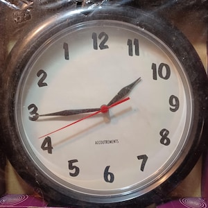 Peut inclure: Horloge ronde avec un cadre noir et un cadran blanc. Les chiffres sont noirs et les aiguilles noires et rouges. Le texte "Backwards Clock" est imprimé au-dessus et en dessous du cadran.