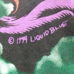 Op de afbeelding: Een zwart T-shirt met een paars en groen ontwerp. De tekst "&copy; 1994 LIQUID BLUE" is op het shirt gedrukt.