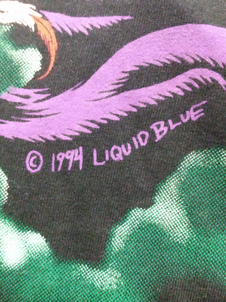 Op de afbeelding: Een zwart T-shirt met een paars en groen ontwerp. De tekst "&copy; 1994 LIQUID BLUE" is op het shirt gedrukt.