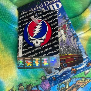 以下が含まれることがあります： グレイトフル・デッドのタグが付いたタイダイTシャツ。タグにはバンドのロゴ、稲妻のボルトが付いたスカル、骸骨の船が描かれています。シャツはカラフルでサイケデリックなデザインです。
