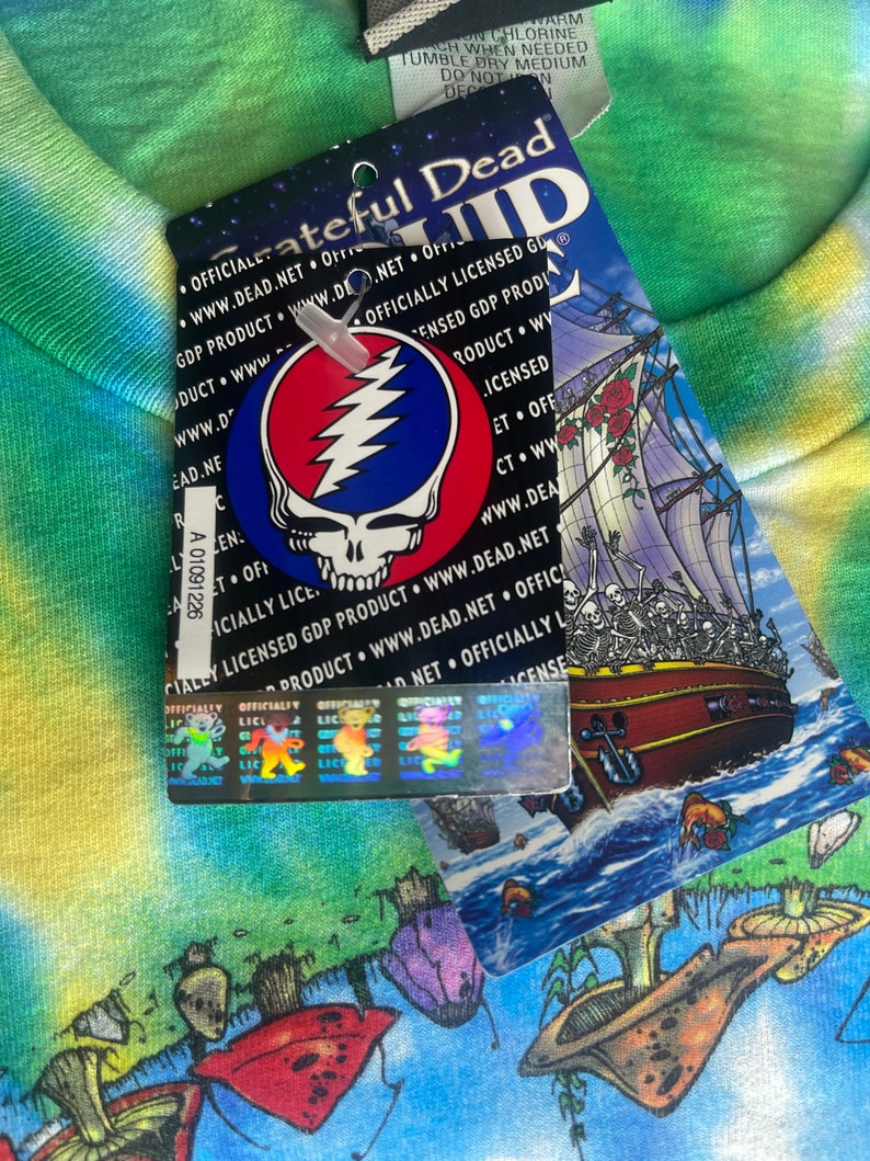 以下が含まれることがあります： グレイトフル・デッドのタグが付いたタイダイTシャツ。タグにはバンドのロゴ、稲妻のボルトが付いたスカル、骸骨の船が描かれています。シャツはカラフルでサイケデリックなデザインです。