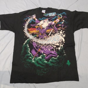 Op de afbeelding: Zwart T-shirt met een grafische print van een tovenaar die een spreuk uitspreekt. De tovenaar draagt een paarse robe en een punthoed. De print is omringd door sterren en wolken. Het T-shirt heeft een Liquid Blue-label.