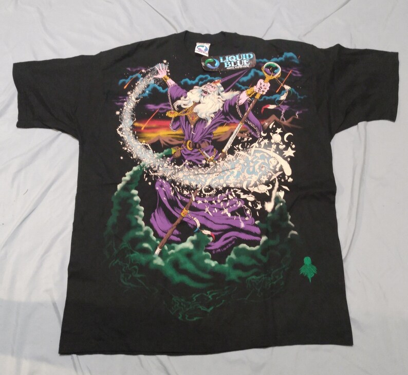 Op de afbeelding: Zwart T-shirt met een grafische print van een tovenaar die een spreuk uitspreekt. De tovenaar draagt een paarse robe en een punthoed. De print is omringd door sterren en wolken. Het T-shirt heeft een Liquid Blue-label.