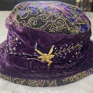 May include: A purple velvet hat with a gold fairy charm and blue bead accents. The hat features an embroidered floral design on the crown and brim. The hat has a wide brim and a cylindrical crown.