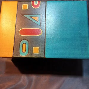 May include: A rectangular wooden box with a teal lid and a decorative front panel. The front panel features geometric shapes in orange, red, and teal against a dark gray background. The box has a shadow underneath.