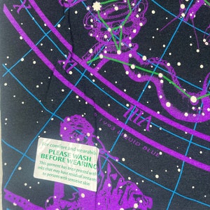 May include: A black t-shirt with a purple and green constellation print. The print includes the text "CRATER" and "1993 LIQUID BLUE". A green tag attached to the shirt reads "PLEASE WASH BEFORE WEARING. This garment has been printed with inks that may have residual irritants to persons with sensitive skin."