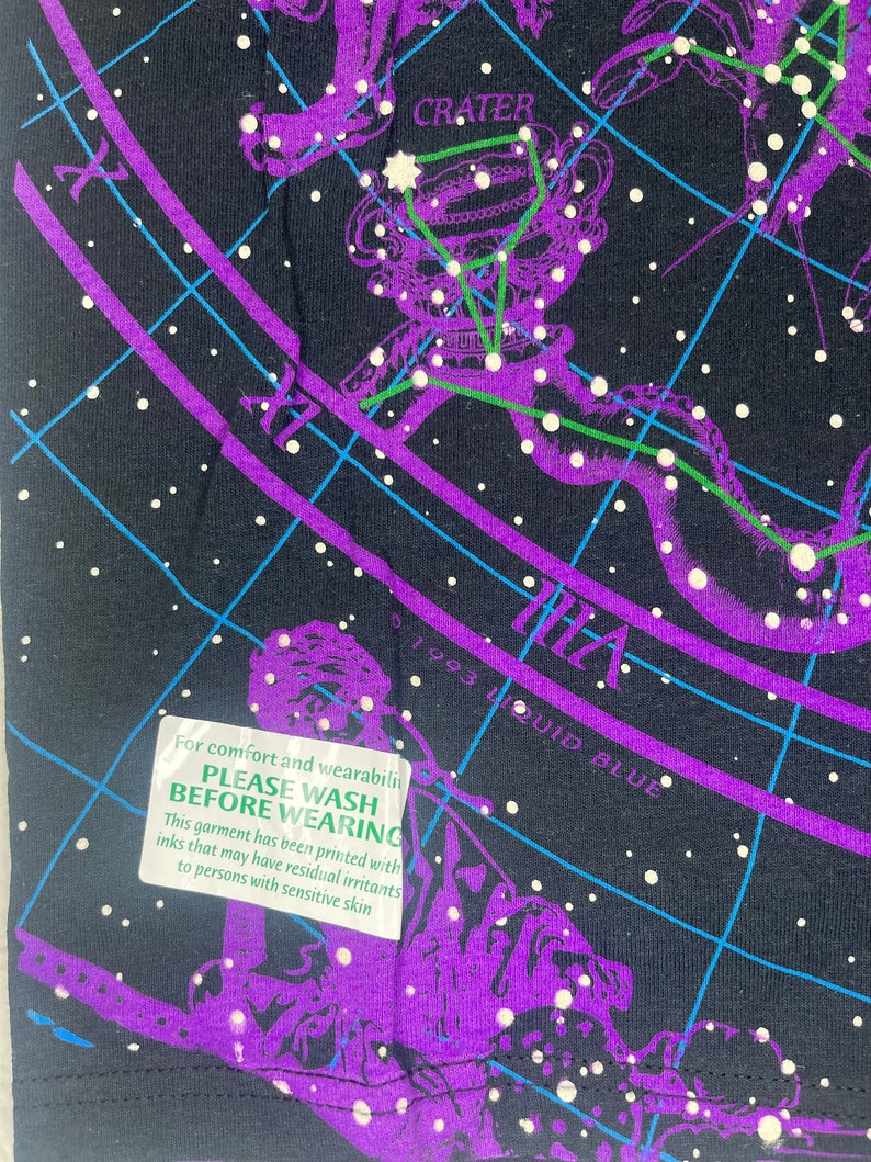 May include: A black t-shirt with a purple and green constellation print. The print includes the text "CRATER" and "1993 LIQUID BLUE". A green tag attached to the shirt reads "PLEASE WASH BEFORE WEARING. This garment has been printed with inks that may have residual irritants to persons with sensitive skin."