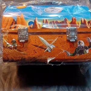 Pode incluir: Uma lancheira de metal com um design temático espacial. A caixa apresenta ilustrações de um foguete, astronautas e uma paisagem marciana com formações rochosas vermelhas sob um céu azul. A lancheira é fixada com dois fechos de metal.