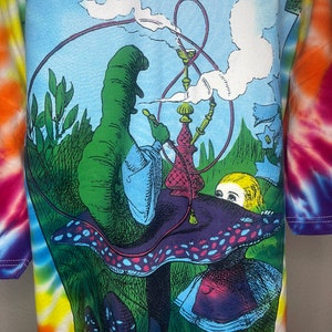 Op de afbeelding: Een tie-dye T-shirt met een kleurrijke illustratie van de rups uit Alice in Wonderland die een waterpijp rookt op een paddenstoel. De rups is groen en blauw, en Alice draagt een blauwe jurk met een rode bovenkant.
