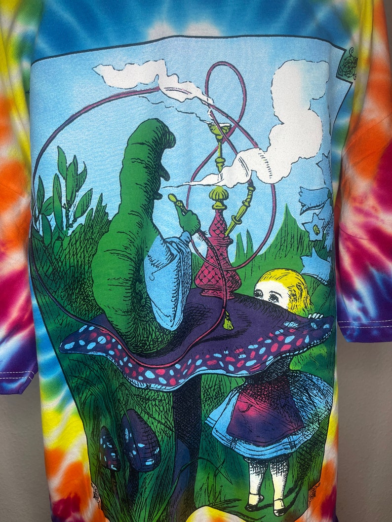 Op de afbeelding: Een tie-dye T-shirt met een kleurrijke illustratie van de rups uit Alice in Wonderland die een waterpijp rookt op een paddenstoel. De rups is groen en blauw, en Alice draagt een blauwe jurk met een rode bovenkant.