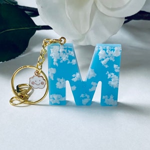 May include: A blue resin letter "M" keychain with a cloud design. The letter is filled with white fluffy clouds, and attached to a gold-colored chain and a charm of a smiling cloud.