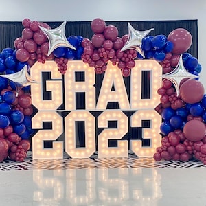 May include: A white light-up sign spelling "GRAD 2023" decorated with maroon and blue balloons and silver star-shaped balloons.