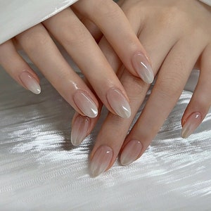 Könnte beinhalten: Nahaufnahme von Händen mit ovalen Nägeln, die mit einem Farbverlauf von Weiß zu Nude lackiert sind.