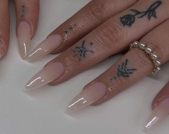 May include: Close-up of long, nude-colored, almond-shaped acrylic nails with a clear, glossy finish. The nails are on a person's hands, which have several small tattoos.