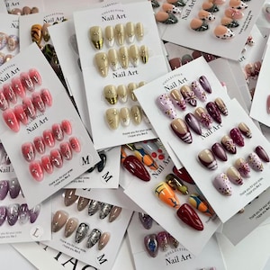 Peut inclure: Collection de faux ongles aux designs et couleurs variés, dont rose, doré et motifs. Chaque ensemble est présenté sur une carte « Nail Art Collection ». L'image présente diverses options de nail art.
