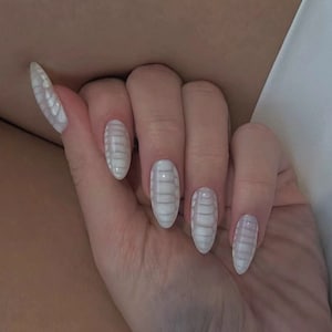 Peut inclure: Une main avec de longs ongles ovales. Les ongles sont peints en blanc avec un motif texturé en forme de reptile dans une nuance plus claire de blanc, créant un effet 3D. Les ongles sont bien manucurés et polis.