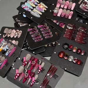 Pode incluir: Uma coleção de vários conjuntos de unhas postiças de pressão exibidas em cartões retangulares pretos. As unhas apresentam diversos designs, incluindo cores sólidas, degradês, acabamentos metálicos e elementos decorativos como strass e berloques.