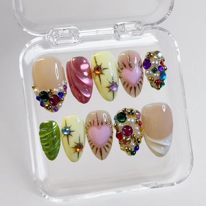 Pode incluir: Um conjunto de dez unhas postiças em uma caixa de plástico transparente. As unhas são decoradas com strass, glitter e detalhes dourados. Algumas unhas têm um design de coração rosa, outras um design de folha verde e outras um design de estrela amarela.