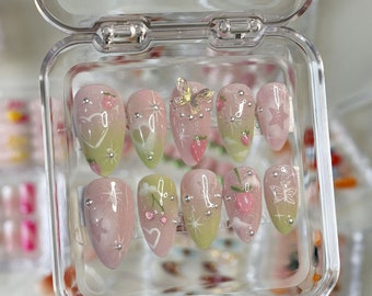 May include: A set of 10 false nails with a variety of pastel pink and green designs. The nails feature butterflies, hearts, stars, and other whimsical details. The nails are displayed in a clear plastic case.
