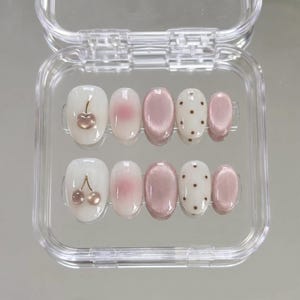 Könnte beinhalten: Ein Set aus zehn künstlichen Fingernägeln in einer durchsichtigen Kunststoffbox. Die Nägel zeigen verschiedene Designs, darunter weiße Basen mit metallischen Kirsch-Applikationen, sanfte rosa Verläufe, metallisches Mauve und weiße Punkte.
