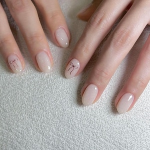 May include: Close-up of hands with oval-shaped nails painted a light beige colour. Two nails have a small black bow design.