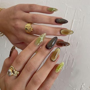 Peut inclure: Gros plan sur une main avec des ongles en amande peints dans un dégradé de nuances de vert et de brun. Certains ongles ont un motif de ligne blanche.