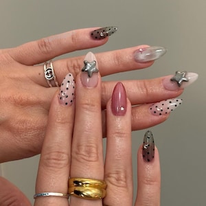 May include: A close-up of manicured nails with various designs. Some nails are clear with black polka dots, others are a mauve color, and some have silver star embellishments. Several rings adorn the fingers.