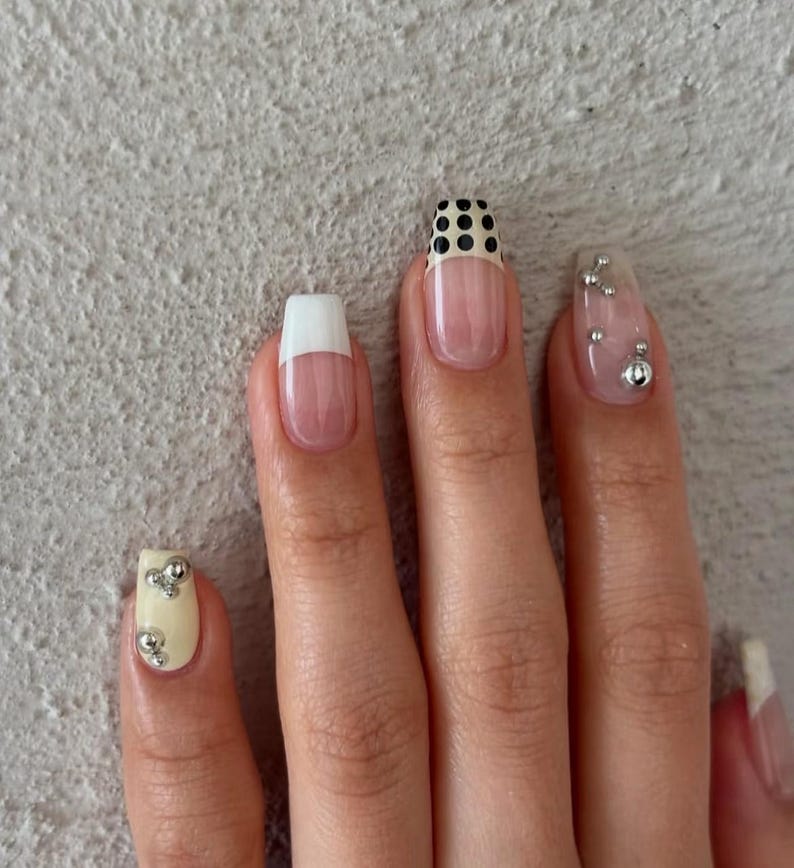 May include: Close-up of a hand with manicured nails. The nails feature various designs, including a white tip, a black and white polka dot pattern, and clear nails with silver embellishments. The nails are a mix of square and oval shapes.
