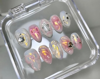 May include: A set of ten iridescent, oval-shaped fake nails with pink and white designs. Each nail has a different design, including a star, a bow, and a swirl. The nails are displayed in a clear plastic case.